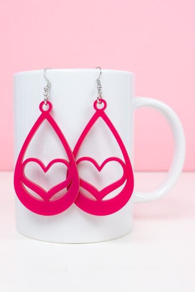 Pink heart earrings hanging on a white coffee cup with a pink background